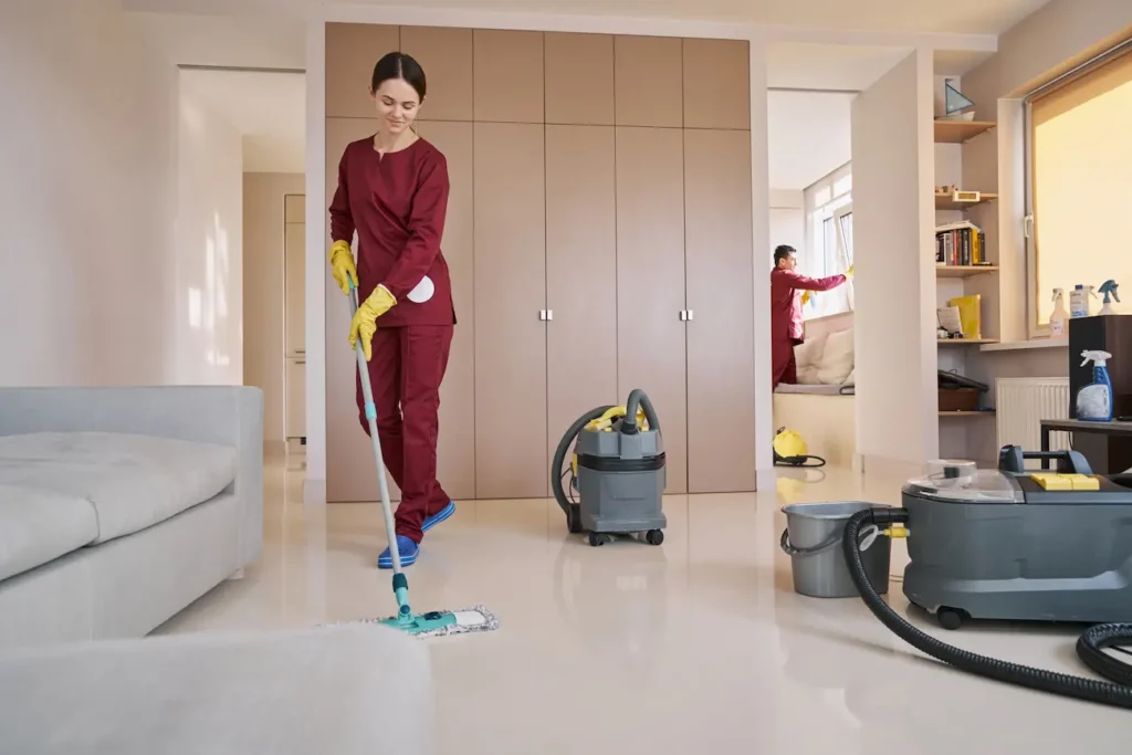 A cleaning professional mops a shiny floor with cleaning equipment and a second worker cleans a window in the background.