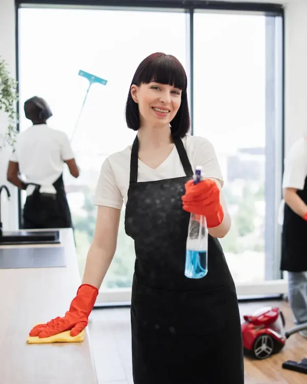 A smiling cleaning professional in gloves holds a spray bottle while a colleague vacuums nearby in a modern office setting.