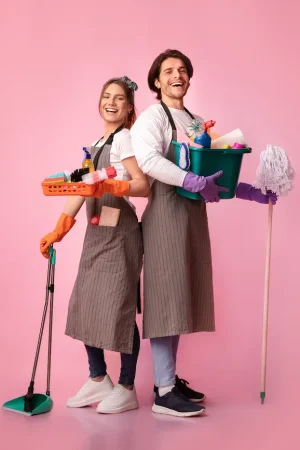 Two smiling cleaners stand back to back against a pink background, holding cleaning supplies and tools for their cleaning