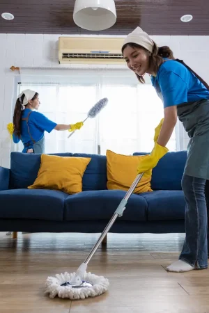 Two women in blue shirts and yellow gloves cleaning a living room, one using a mop and the other dusting.
